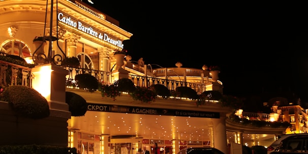 A luxurious casino at night, illuminated with warm, golden lights. The building features elegant architecture with columns and decorative elements. A few people are walking towards the entrance, and there are a couple of parked cars nearby.