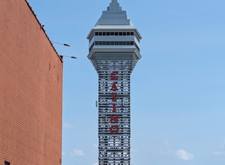 A tall tower with a modern design features the large vertical red letters spelling 'CASINO'. The structure has a sleek, metallic appearance with various horizontal and vertical lines. The sky is mostly clear with a few clouds. In the foreground, part of a brick building is visible, along with some trees and buildings in the distance.