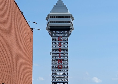 A tall tower with a modern design features the large vertical red letters spelling 'CASINO'. The structure has a sleek, metallic appearance with various horizontal and vertical lines. The sky is mostly clear with a few clouds. In the foreground, part of a brick building is visible, along with some trees and buildings in the distance.