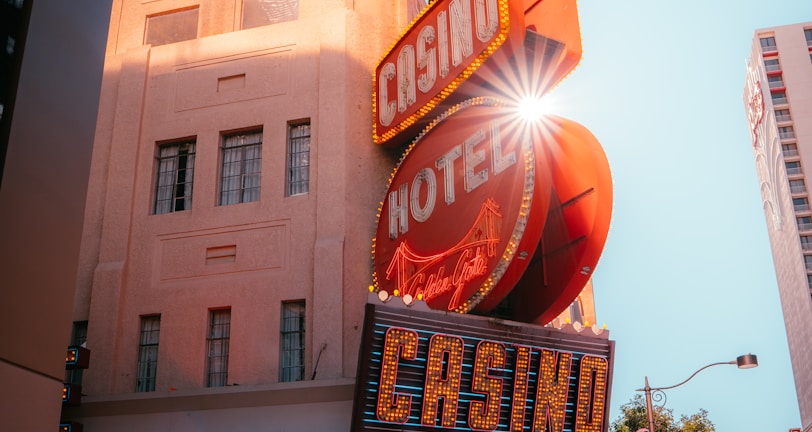 A vibrant city scene featuring a tall, illuminated casino sign with retro-style neon lights. The sign dominates the building facade with the words 'CASINO' and 'HOTEL'. Sunlight peeks through, creating a dramatic lens flare effect. The street is lively, with pedestrians casually walking and a hint of urban hustle.